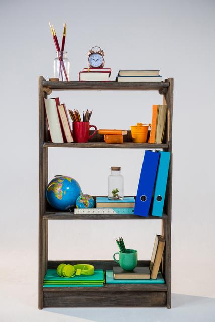 This image shows a neatly organized bookshelf with various office supplies and decorative items. It includes books, binders, a globe, a clock, and stationery such as pencils and pens. Ideal for illustrating concepts of organization, productivity, and workspace setup. Suitable for use in articles or advertisements related to home office organization, study tips, or interior decor.