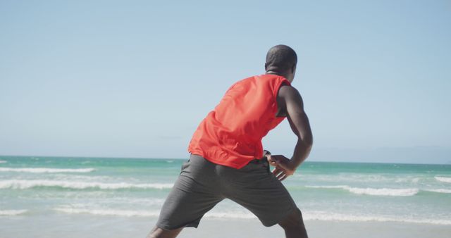 Man wearing red shirt and gray shorts stretching by the ocean on a sunny day. Perfect for workout magazines, fitness blogs, healthy living advertisements, and vacation promotions.