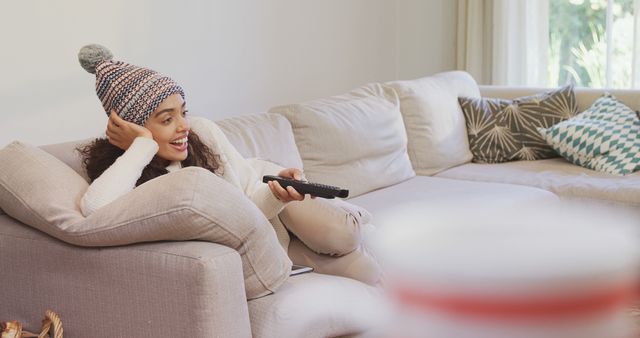 A young African American woman is relaxing on a sofa, wearing a beanie and holding a remote control, with copy space. She appears to be enjoying some leisure time, watching television or selecting a movie to watch.
