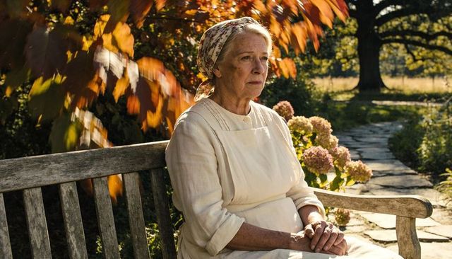 Senior woman sitting on wooden bench wearing apron and floral headscarf in autumn garden