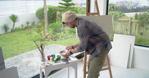 Indian artist preparing paints and mixing media in bright coastal studio with easel