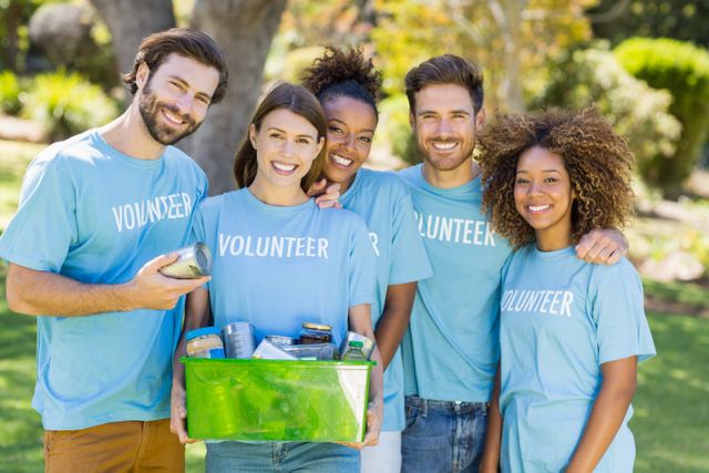 Diverse group of volunteers holding a donation box filled with canned goods in a park. They are smiling and wearing matching blue volunteer shirts. Ideal for use in campaigns promoting community service, charity events, teamwork, and social work. Perfect for illustrating concepts of unity, support, and humanitarian efforts.