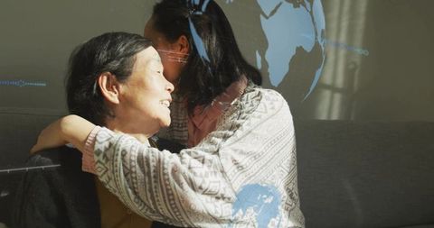 Daughter hugging elderly mother on sofa with blue globe projection, showing warmth