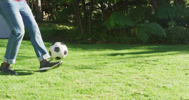 An individual in casual attire, notably jeans and sneakers, is captured in motion, kicking a soccer ball on a lush green lawn. The sunlit surroundings hint at a pleasant, outdoor setting, likely a garden or backyard. Perfect for use in advertisements related to sports equipment, outdoor activities, fitness promotions, or casual clothing.