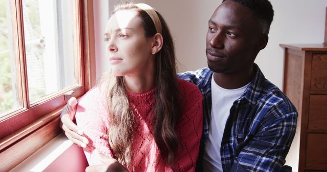 Interracial couple sitting near a window, reflecting together. Ideal for themes on relationships, diversity, and emotional connection. Suitable for use in articles, blogs, and advertisements about love, inclusivity, and personal moments.