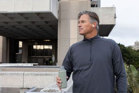 Middle-aged man on urban plaza holding textured bottle wearing earbuds and quarter-zip
