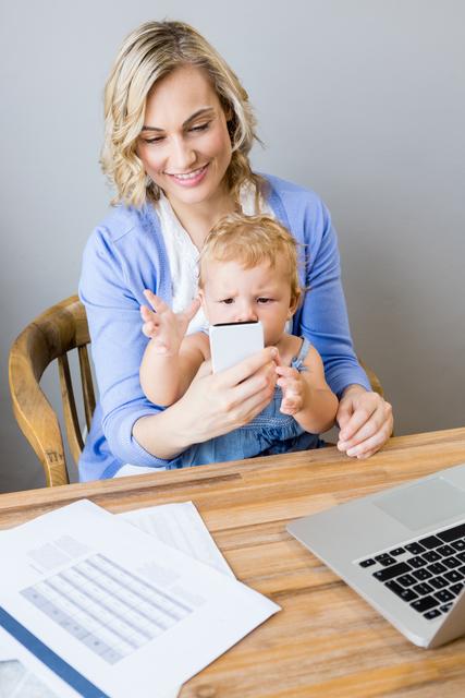 Mother and baby sitting at table and using mobile phone at home