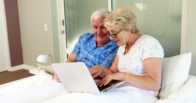 Senior couple sitting up in bed using laptops, demonstrating technology use among the elderly. Bright and relaxed bedroom environment. Perfect for topics on seniors' technology adoption, retirement lifestyle, staying connected, or aging and digital literacy.