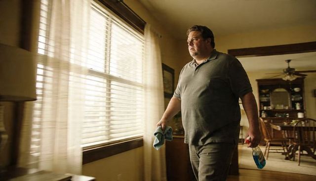 Overweight man cleaning home, walking with spray bottle and cloth in sunlit living room