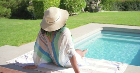 African american woman relaxing by backyard pool wearing straw hat and striped cover-up