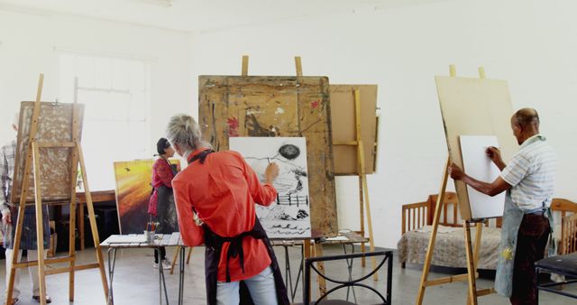 Group of artists actively engaged in a creative art workshop, painting and drawing at easels in a well-lit studio. Useful for illustrating creativity, adult education, and collaborative artistic environments.