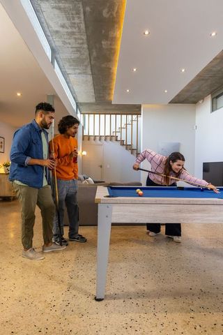 Friends playing pool in modern lounge with concrete ceiling and blue felt table, drinks