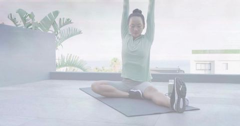 Seated straddle stretching on rooftop terrace with tropical plants for morning wellness