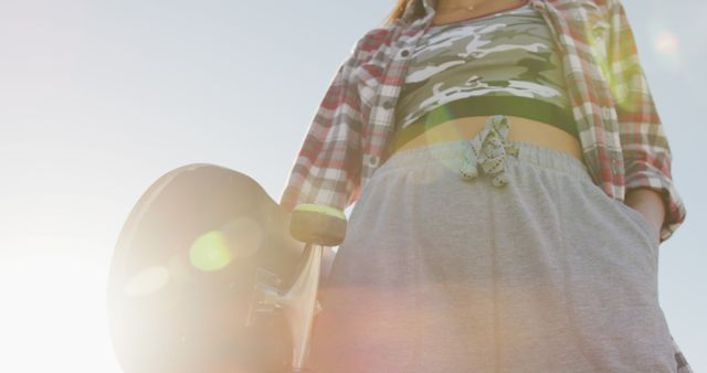 This image shows a young woman holding a skateboard while enjoying a summer day. She is dressed in a casual plaid shirt and loose trousers. It can be used for themes related to youth culture, outdoor recreational activities, summer fun, and urban lifestyles.