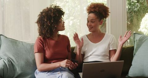 Female friends chatting on couch with laptop and smartphone, planning together at home