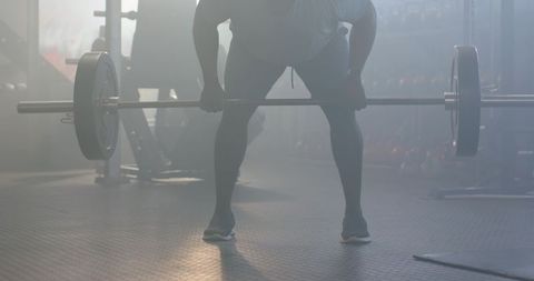Backlit male athlete preparing barbell on rubber gym floor with mist silhouette