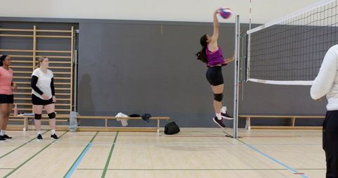 Asian player jumping and spiking volleyball over net while teammates watching indoor court