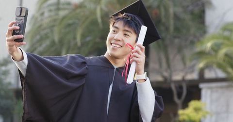 Asian graduate taking selfie wearing cap and gown holding diploma on campus
