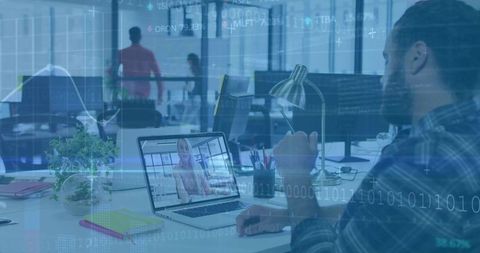 Bearded man watching remote video call on laptop in open office with data visualization