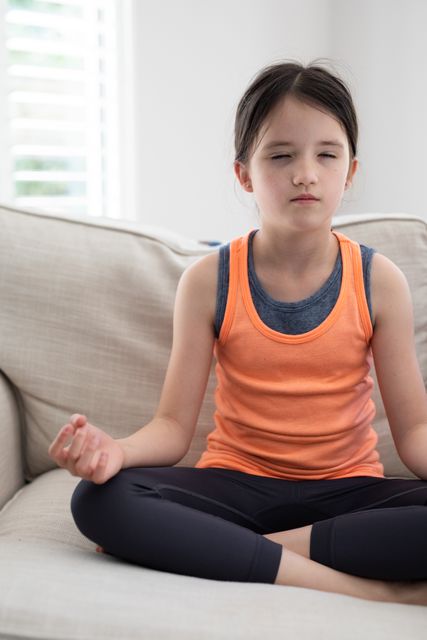 Young girl sitting on a couch with legs crossed, meditating with eyes closed. She is wearing an orange tank top and appears to be in a state of relaxation and mindfulness. This image can be used for promoting mental health, wellness, and mindfulness practices for children. It is also suitable for educational materials, parenting blogs, and lifestyle articles focusing on relaxation and tranquility.