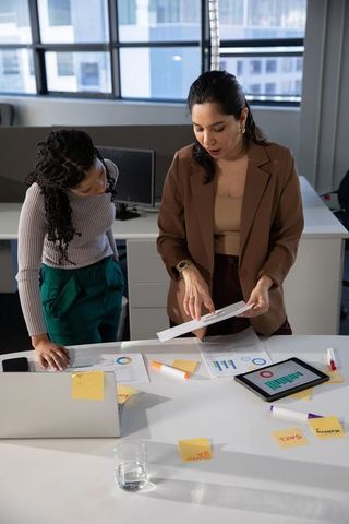 Diverse Women Collaborating Over Charts, Tablet and Laptop in Modern Office Natural Light