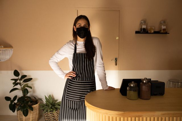 Female cafe owner wearing a face mask and apron, leaning on the counter in a modern cafe. The setting includes indoor plants and jars, reflecting a clean and welcoming environment. Ideal for use in articles or advertisements about small businesses, entrepreneurship, health and safety measures during the pandemic, or the hospitality industry.