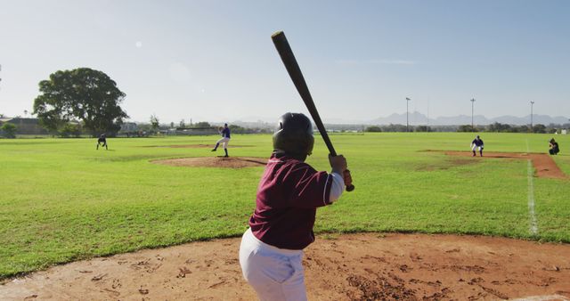 Boy in batting stance preparing to hit ball during sunny practice game. Teammates playing on field with green grass and blue sky. Great for articles, sports training materials, and summer camp promotions.