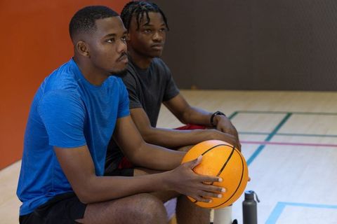 Two african american teammates sitting on bench holding basketball on indoor court