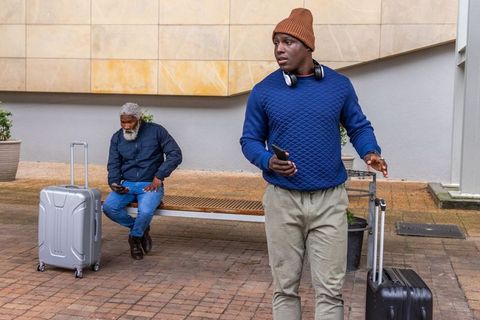 Young man texting while pulling suitcase past seated traveler in urban transit plaza
