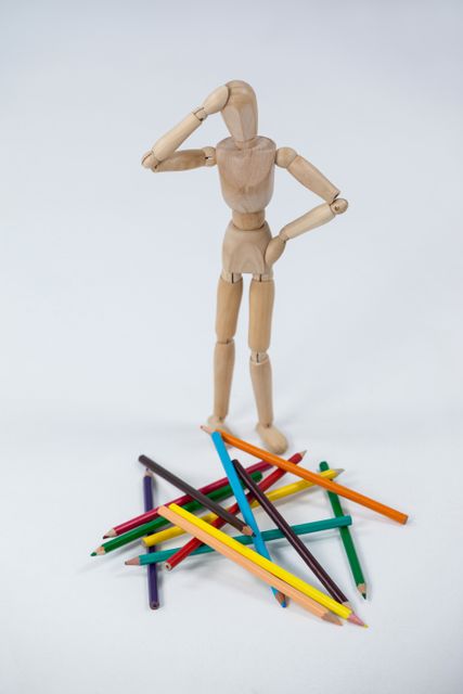 Confused wooden figurine standing near a heap of color pencils against white background