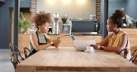 Two african women collaborating over laptop and papers in rustic coffee shop planning