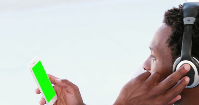 Man wearing headphones checking a green screen smartphone while listening to music or podcast. Perfect for technology, music streaming, mobile app presentation, and social media content demonstrating device functionality or promoting audio services.