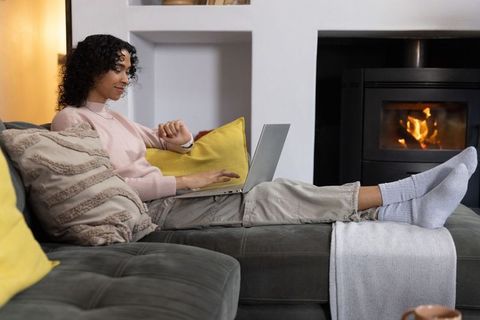 Non-binary person reclining on sofa using laptop and checking watch by fireplace