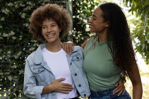 African american women embracing and smiling in sunny garden wearing denim jacket and green top