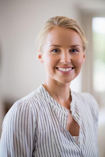 Blonde woman smiling warmly while standing indoors, wearing a striped shirt. Ideal for use in lifestyle blogs, home decor advertisements, and promotional materials emphasizing friendliness and approachability.