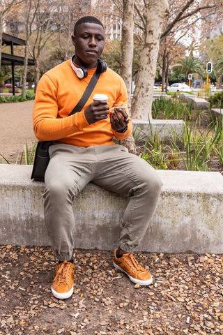Sitting man in orange sweatshirt holding coffee and pastry at urban plaza wearing headphones