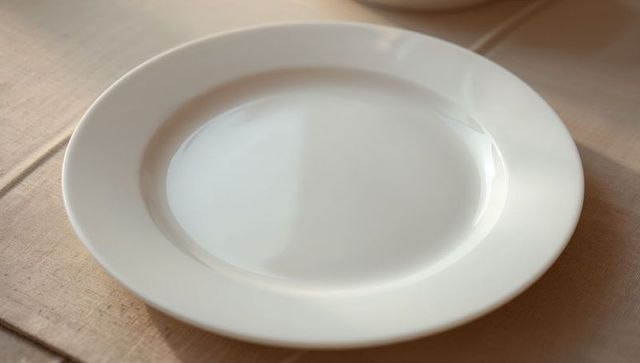 Minimalist white ceramic plate sitting on beige woven tablecloth with soft warm light