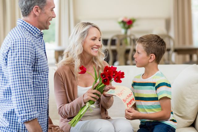 Boy presenting card and roses to his mother in a cozy living room. Perfect for themes of family bonding, Mother's Day, celebrations, and expressions of love. Ideal for use in advertisements, greeting cards, and parenting blogs.