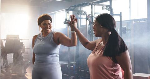 Diverse women high-fiving after strength training in sunlit gym for inclusive fitness