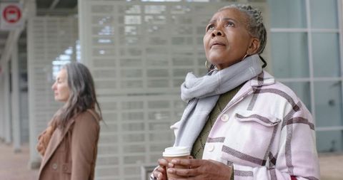 Senior african american woman holding coffee while waiting at urban bus stop, contemplative gaze