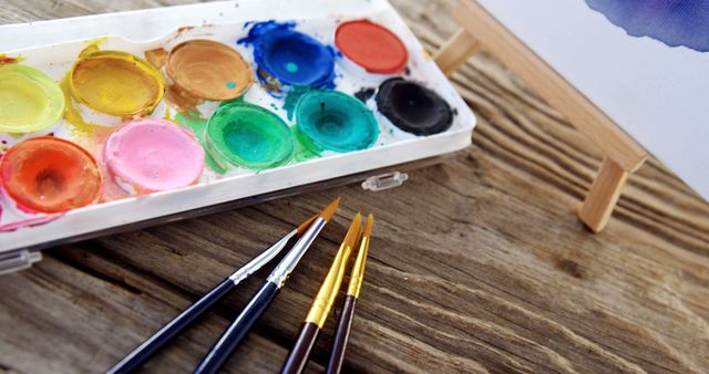 A well-used watercolor palette with vibrant colors lies next to brushes on a wooden surface, with copy space. Art supplies like these invite creativity and are essential for artists to express their vision on canvas.