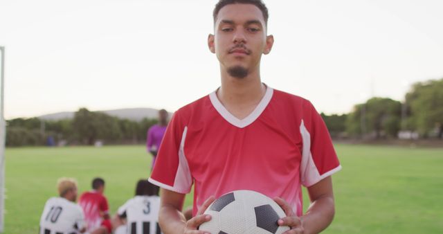Young male soccer player wearing red jersey holding a soccer ball on a grassy field with teammates, dressed in black-and-white jerseys, sitting in the background. Perfect for illustrating sports team dynamics, youth sports activities, team spirit, outdoor training sessions, and athletic lifestyle.