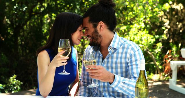 A young Caucasian couple enjoys a romantic moment with a toast in a lush garden setting, with copy space. Their affectionate gaze and the outdoor dining setup suggest a celebration or intimate date.