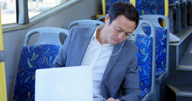 Image depicts a businessman dressed in a suit traveling on a bus while engaged with his laptop and listening through earphones. This visual is ideal for illustrating themes related to commuting, remote work, and modern professional life. Suitable for content related to transportation, business travel, productivity, and city living.