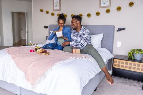 Happy black couple opening gifts on cozy bed with romantic breakfast, pink throw, garland decor