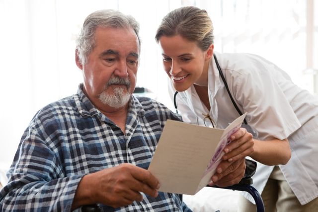 Female doctor showing greeting card to elderly patient sitting in wheelchair in nursing home. Ideal for use in healthcare, senior care, and medical professional contexts, highlighting patient interaction and caregiving.