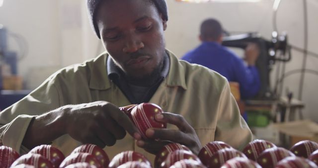 This image shows a man in a factory setting carefully inspecting red sports balls, indicating quality control during the manufacturing process. Useful for illustrating articles about industrial work, manufacturing processes, quality assurance, and labor-intensive jobs. Ideal for topics related to factory work, craftsmanship, and sports equipment production.