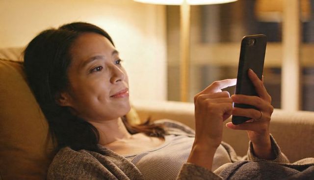 Asian woman reclining on sofa late-night scrolling smartphone under warm lamp glow