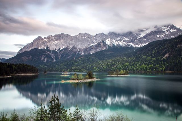 Depicts a tranquil lake with calm waters reflecting surrounding rocky mountains and cloudy sky. Ideal for use in travel brochures, nature magazines, websites or posters promoting outdoor activities, tourism, scenic beauty, and tranquility. Suitable for desktop wallpapers and social media posts on nature and relaxation.