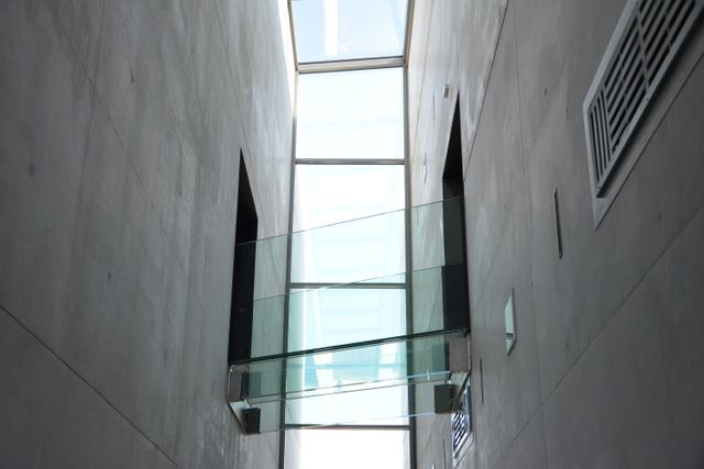 An image of a modern glass skybridge connecting two concrete walls. The minimalist design emphasizes clean lines and geometric shapes, with natural light filtering through a glass skylight above. Ideal for architectural publications, design inspiration, urban planning projects, and contemporary building concepts.